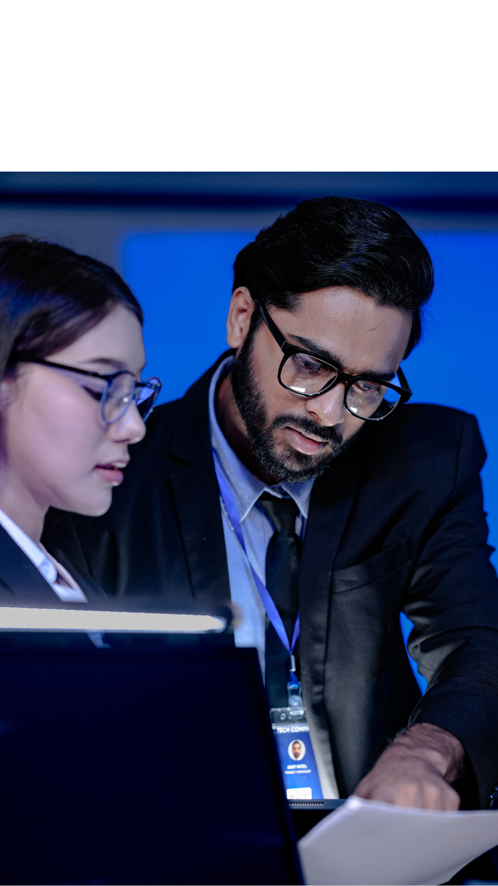 Two professionals reviewing a document beside computer monitors in a blue-lit technology workspace.